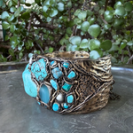 Wide handmade turquoise cuff bracelet featuring large blue-green stones, pearl accents, intricate antique bronze-tone sculptural detailing and safety chain.