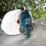 Handmade Blue Apatite pendant necklace combining raw and polished blue gemstones with contrasting textures in an ornate antique bronze-tone polymer clay setting on a black cord.
